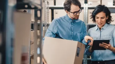 Two people in a warehouse checking a tablet and holding a box.