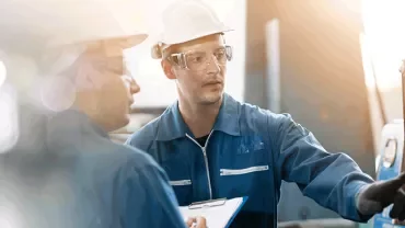 Two workers in protective gear discuss machine operations in a factory setting.