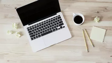 A laptop, coffee cup, pencils, and crumpled papers on a wooden desk.